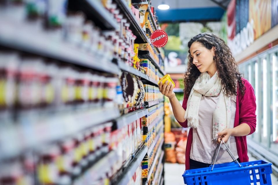 Une femme examine un produit dans un rayon de magasin en tenant un panier bleu.